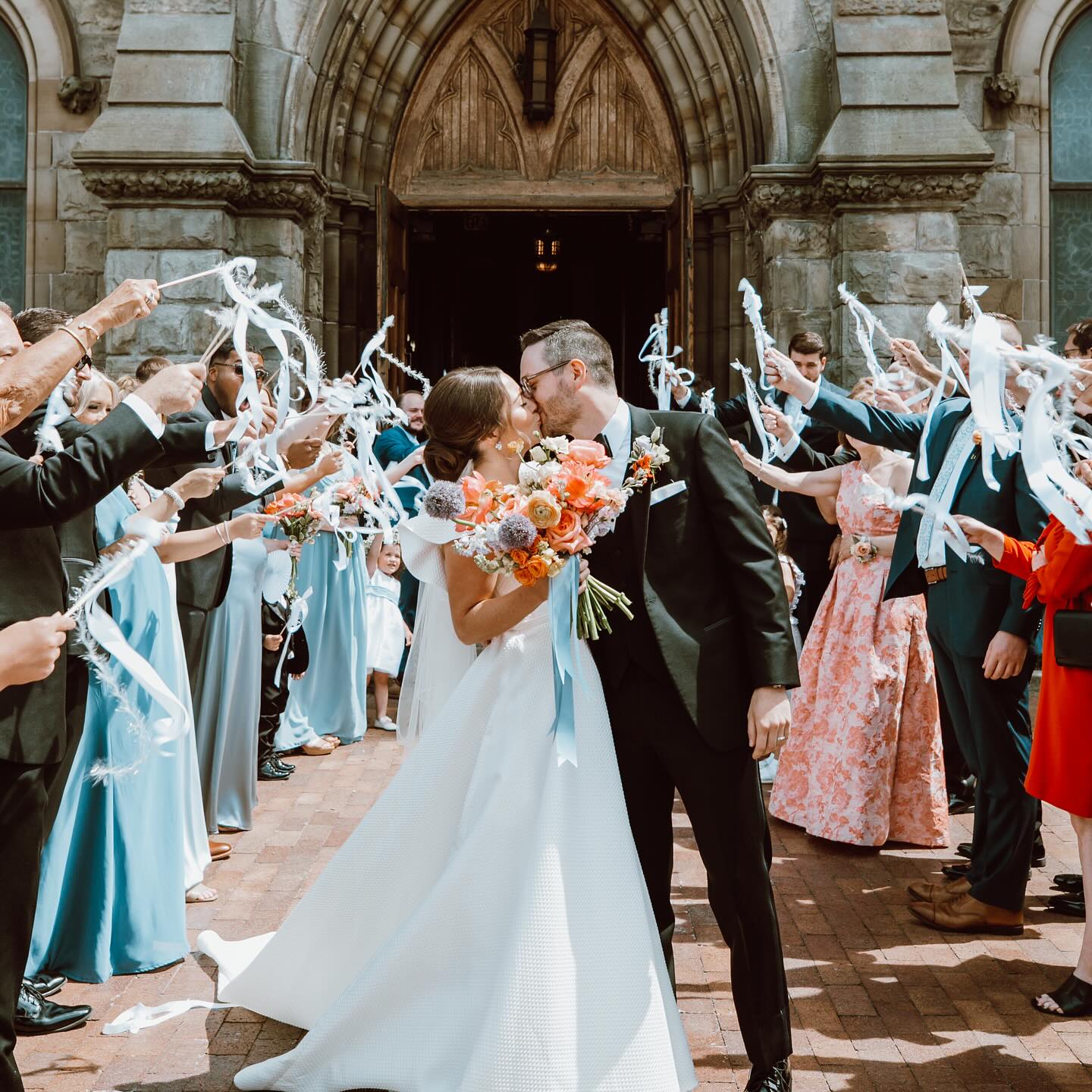 Newlyweds kiss outside a church as guests wave ribbons, bride holding affordable Chicago wedding bouquet.