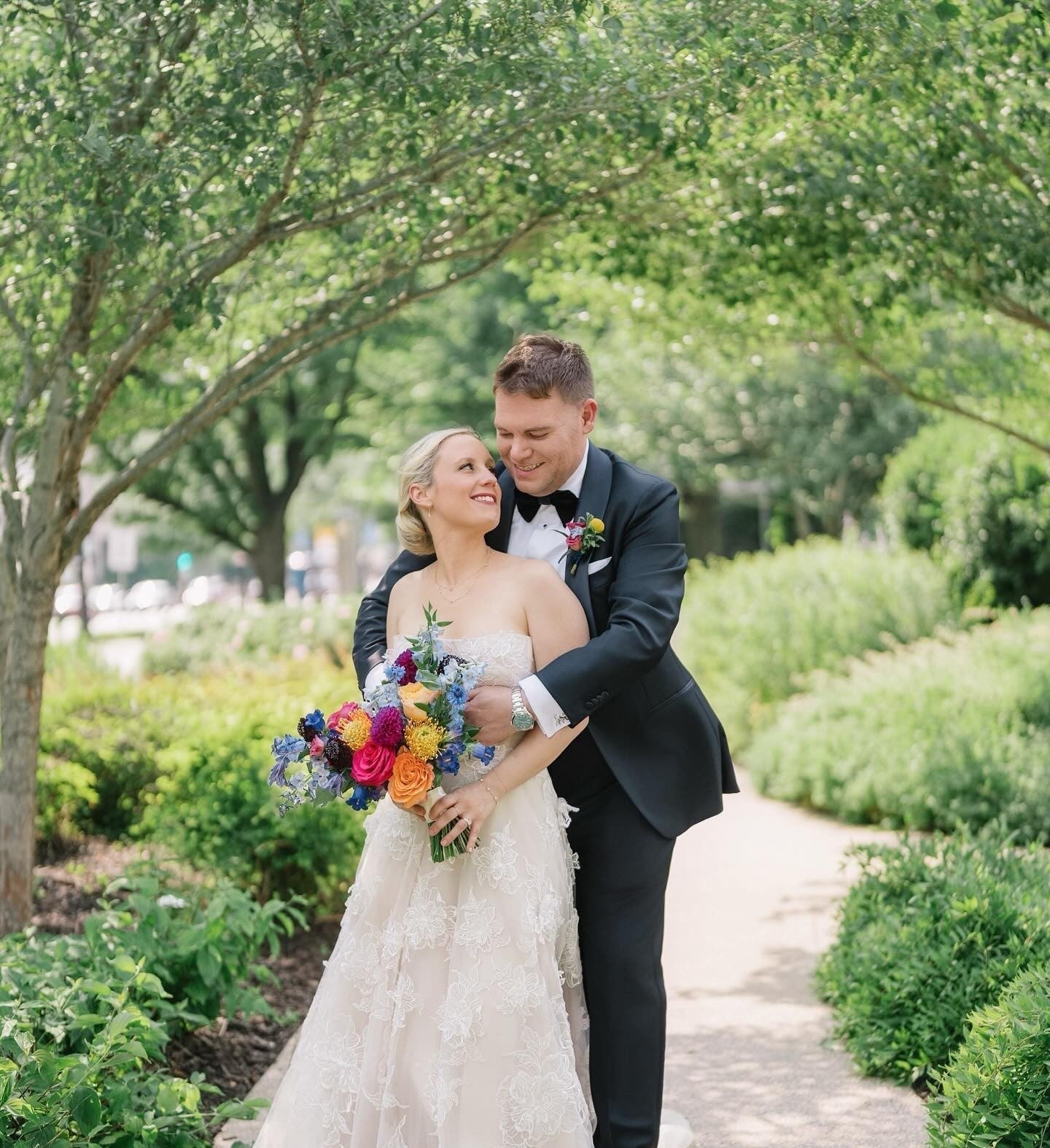 Bride in a lace gown holding a colorful Flowers for Dreams bouquet smiles at the groom in a black suit on a lush garden path.