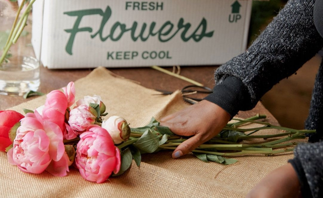 Florist wraps vibrant pink peonies in burlap beside a box labeled "Fresh Flowers: Keep Cool."