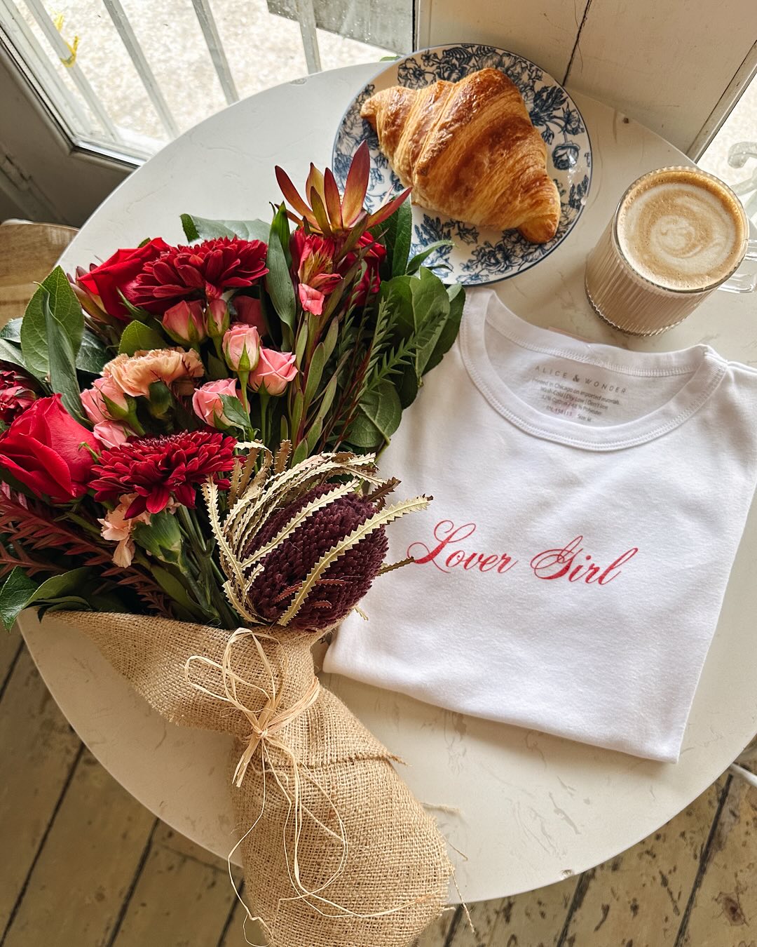  Galentine’s Day pairing of coffee, croissant, Alice & Wonder “Lover Girl” tee with red flower arrangement by Flowers for Dreams.