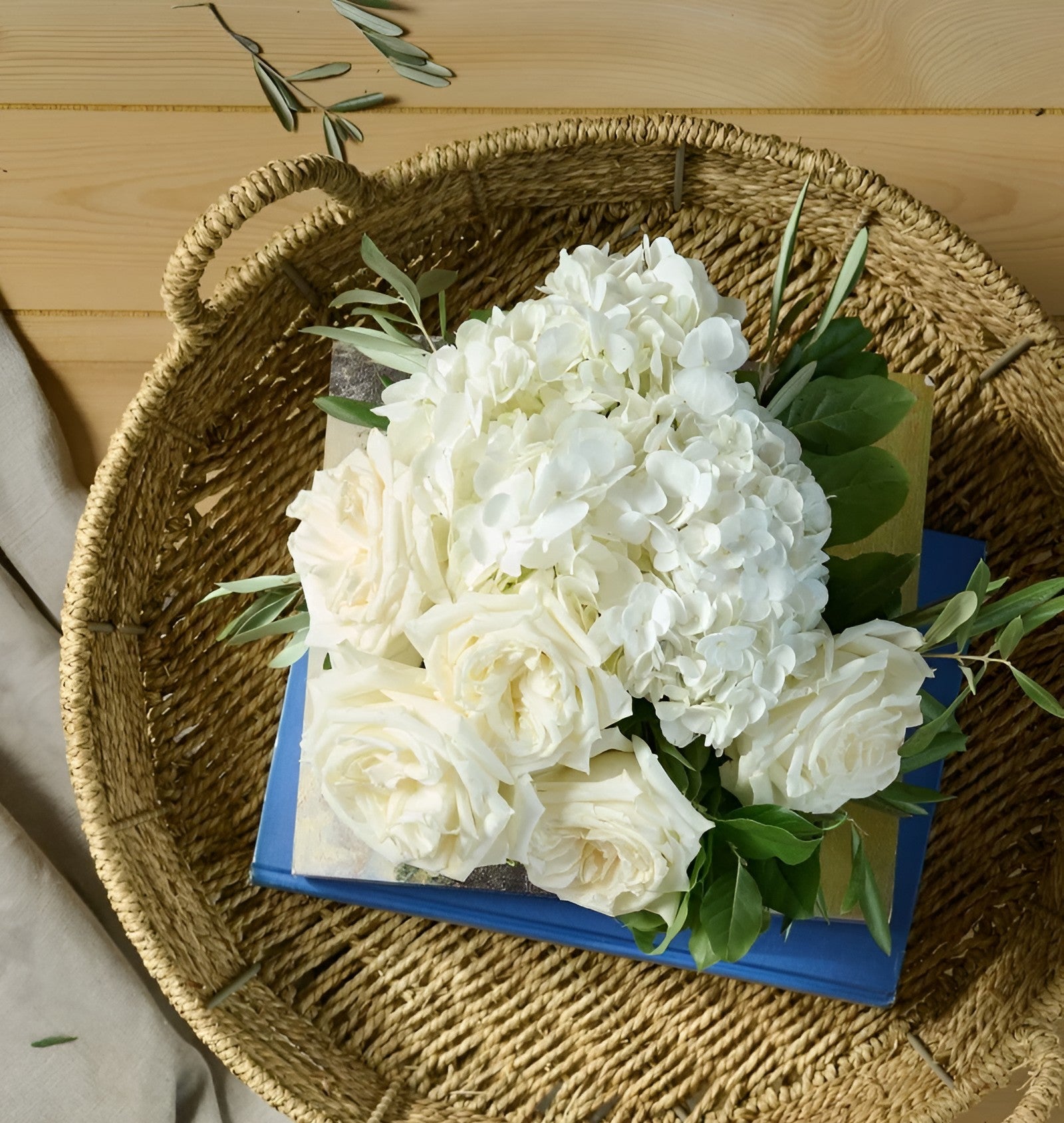  Flower for Dreams white rose and hydrangea sympathy bouquet rests on blue books in a woven basket.
