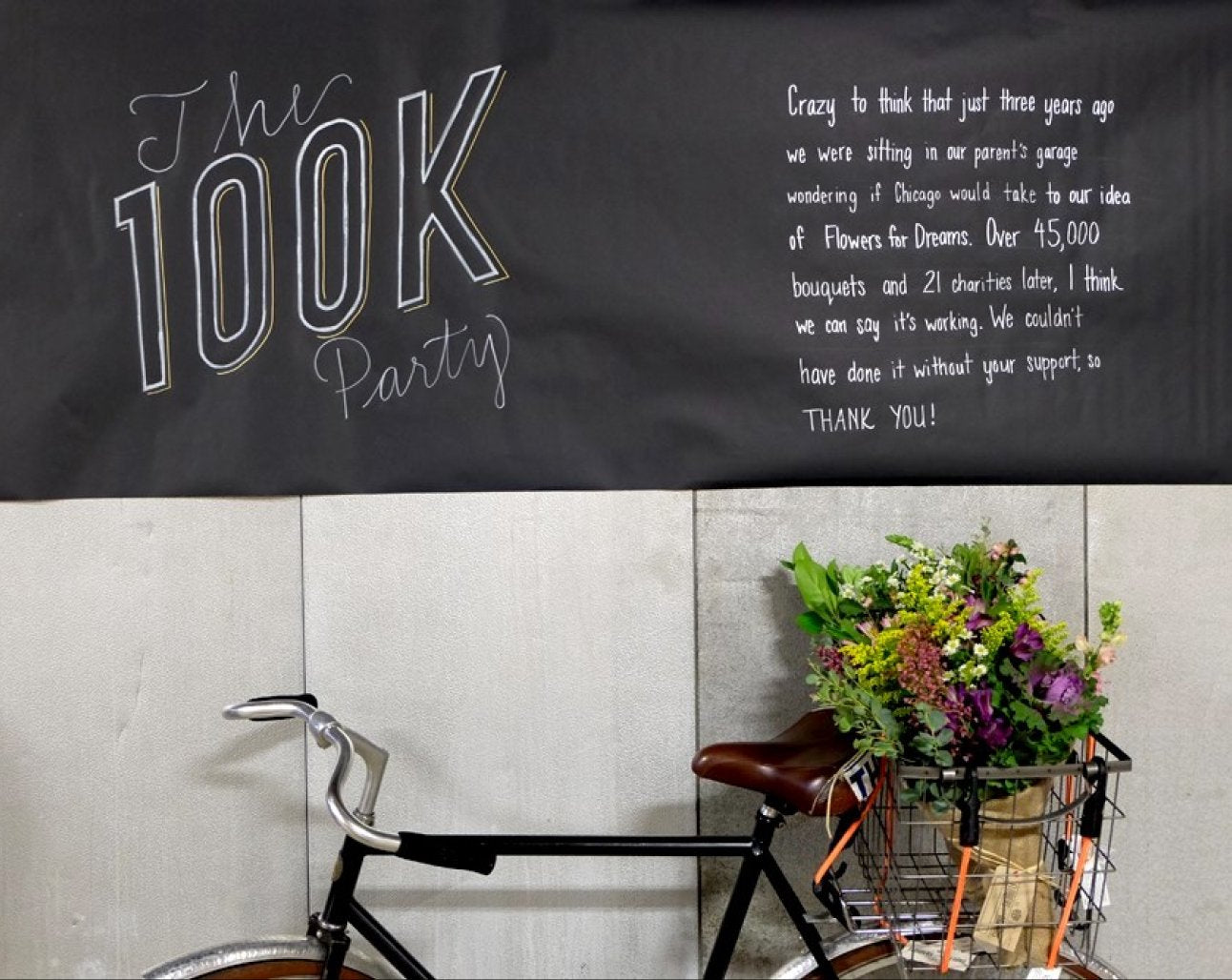 Black banner with white text celebrating a 100K donated milestone, bicycle, and flowers in a basket against a light-colored wall.