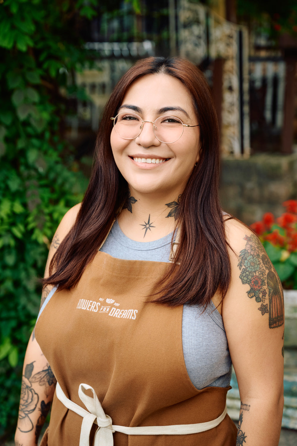 Woman wearing a brown apron with visible tattoos, standing outdoors.