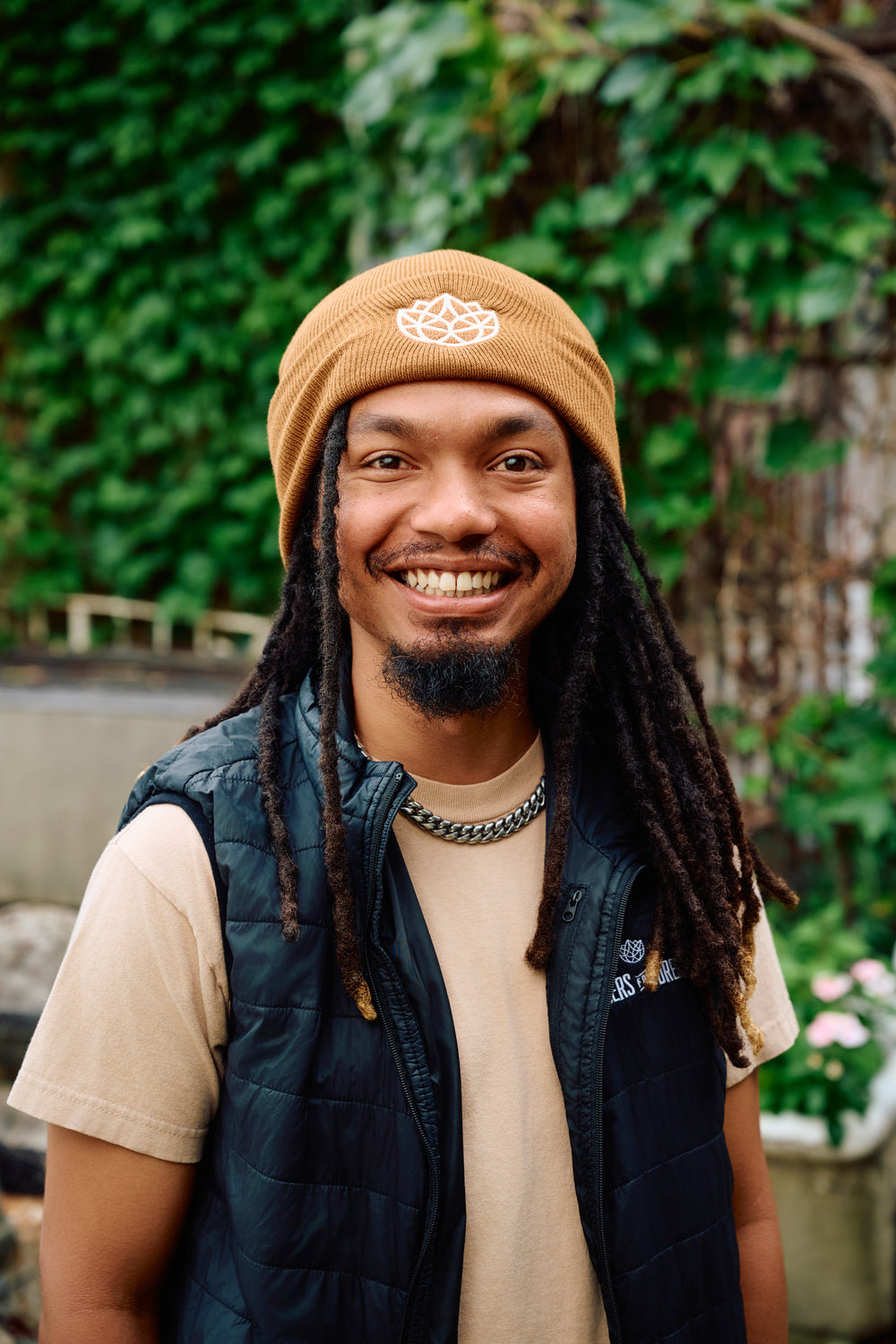 Man wearing a brown beanie and black vest with greenery in the background