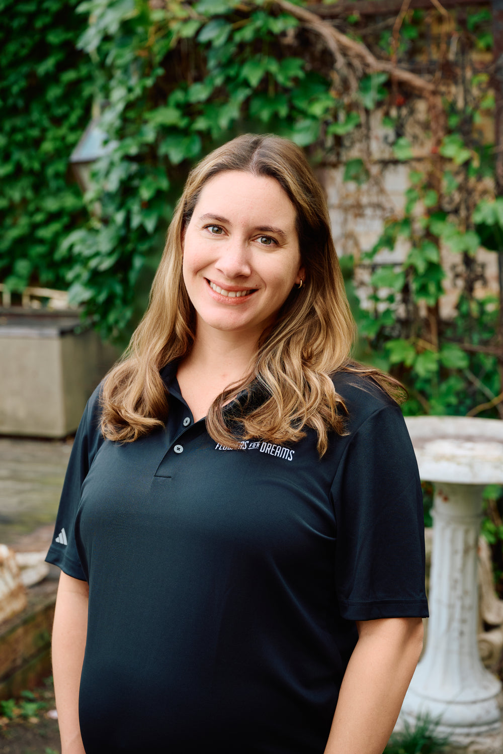 Woman wearing a dark green polo shirt with a logo, standing outdoors against a green leafy background.