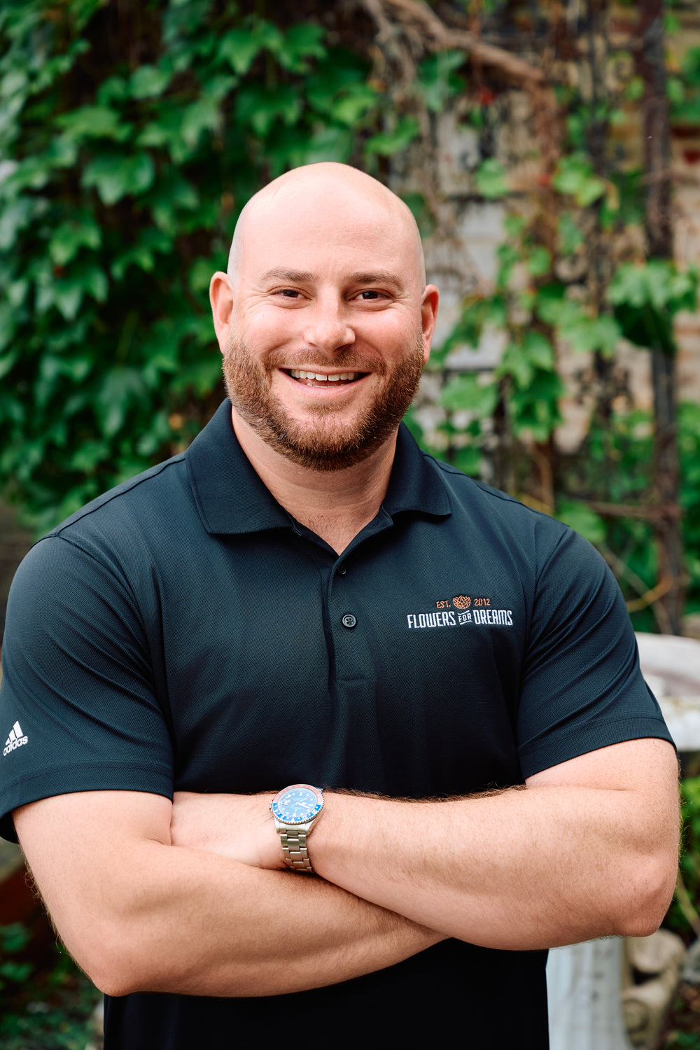 Man wearing a dark blue polo shirt with logos, standing outdoors with greenery in the background
