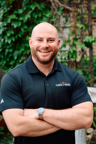 Man wearing a dark blue polo shirt with logos, standing outdoors with greenery in the background