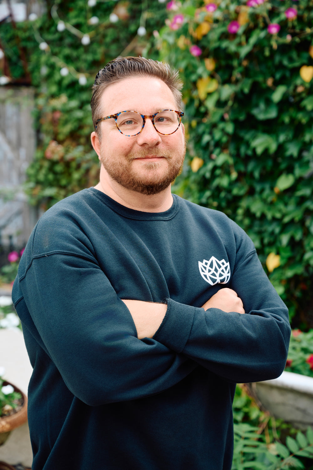 Man wearing a dark green sweatshirt with a logo, standing outdoors with greenery in the background