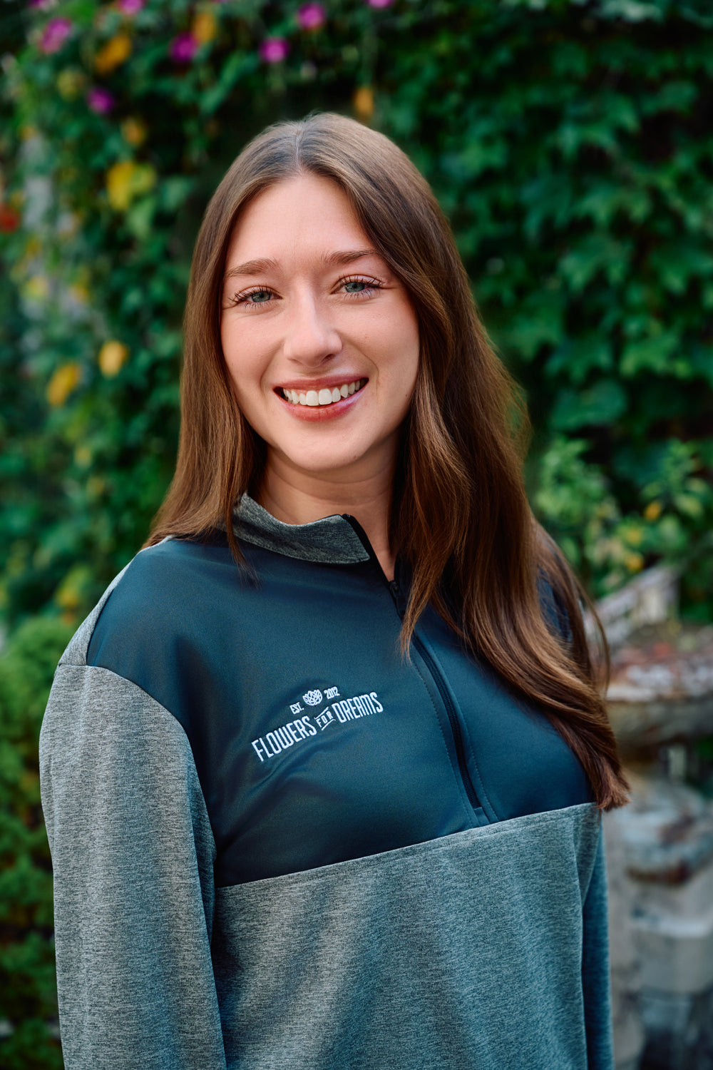 Woman wearing a blue and gray jacket with 'Flowers for Dreams' logo, standing in front of green foliage.
