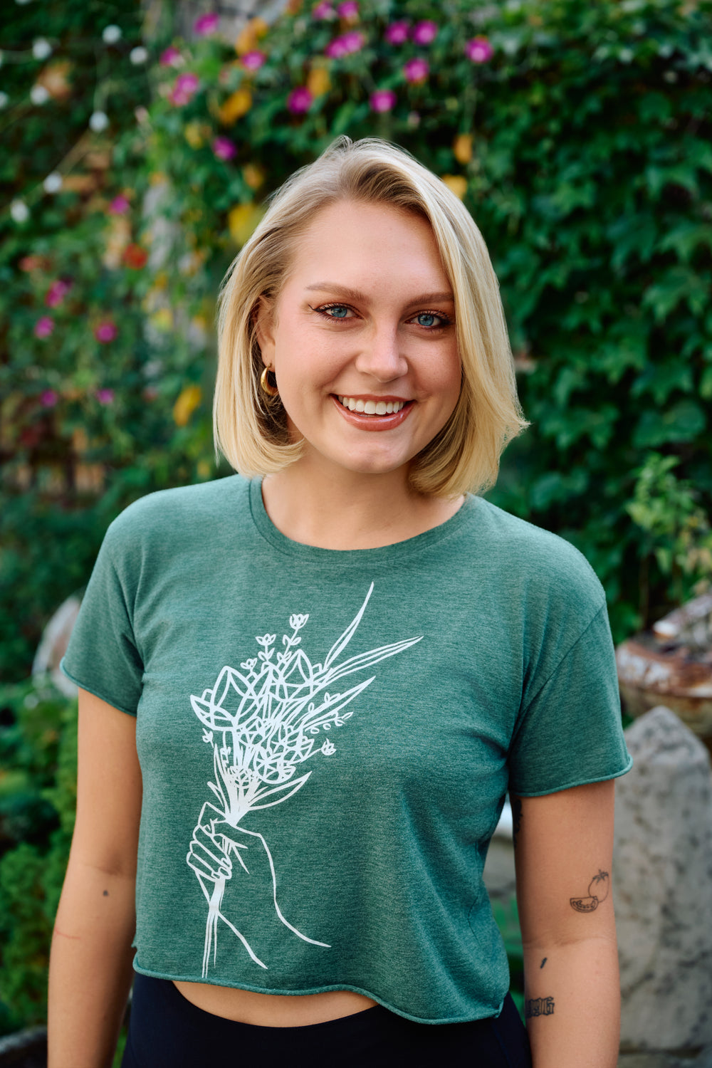 Woman wearing a green t-shirt with a floral design, standing outdoors with greenery in the background.