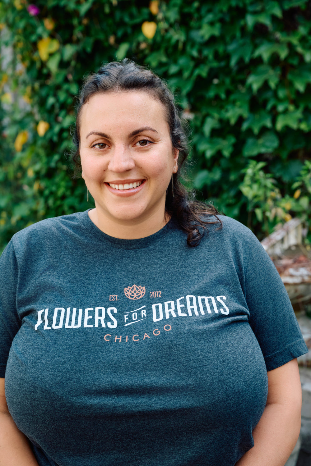 Person wearing a 'Flowers for Dreams Chicago' t-shirt in front of green foliage.