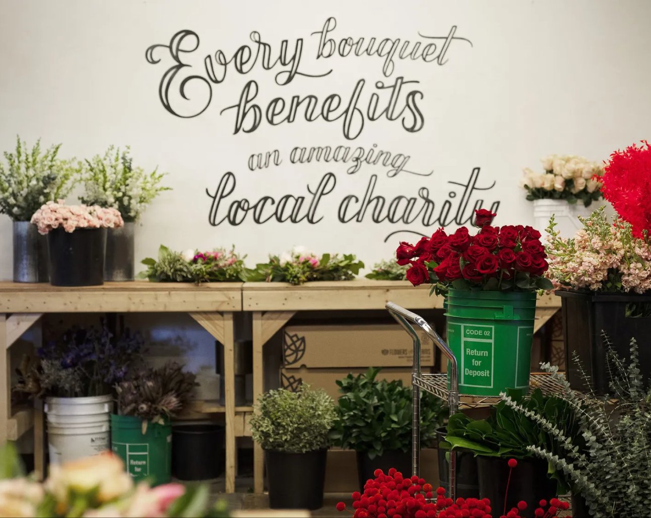 Floral arrangement workshop with text 'Every bouquet benefits an amazing local charity' on a white wall.