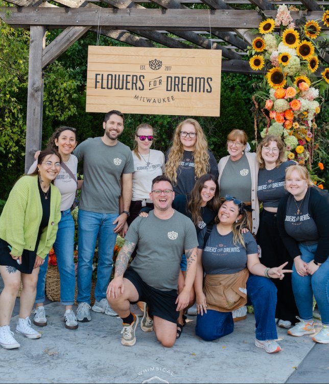 Group of employees posing in front of 'Flowers for Dreams' sign with floral decorations.