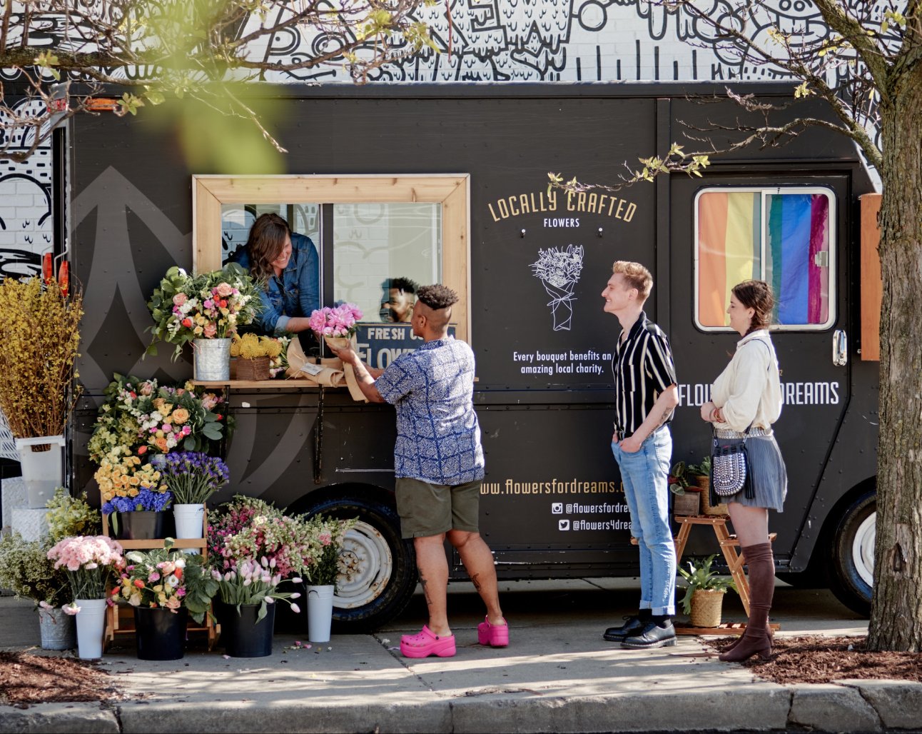 Floral truck with people interacting, surrounded by potted flowers on a street.